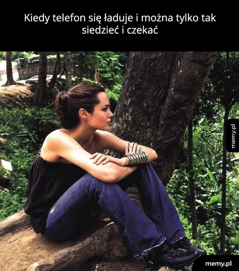 Kobieta siedzi na kamieniu w lesie, czekając na naładowanie telefonu.